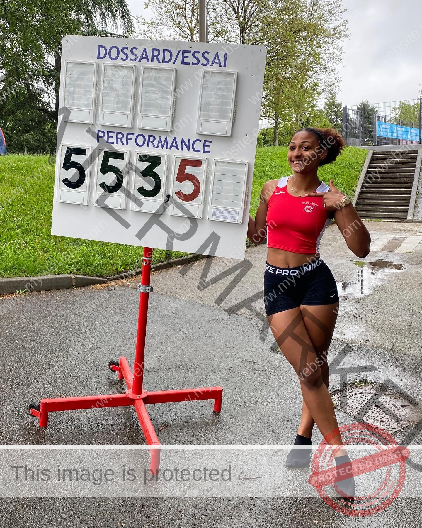 zola-ndouma-mona-france-zolaathle-09444 Zola Ndouma Mona, track and field star from France smiles in sportswear by a scoreboard showing 5.53 outdoors on a rainy day.