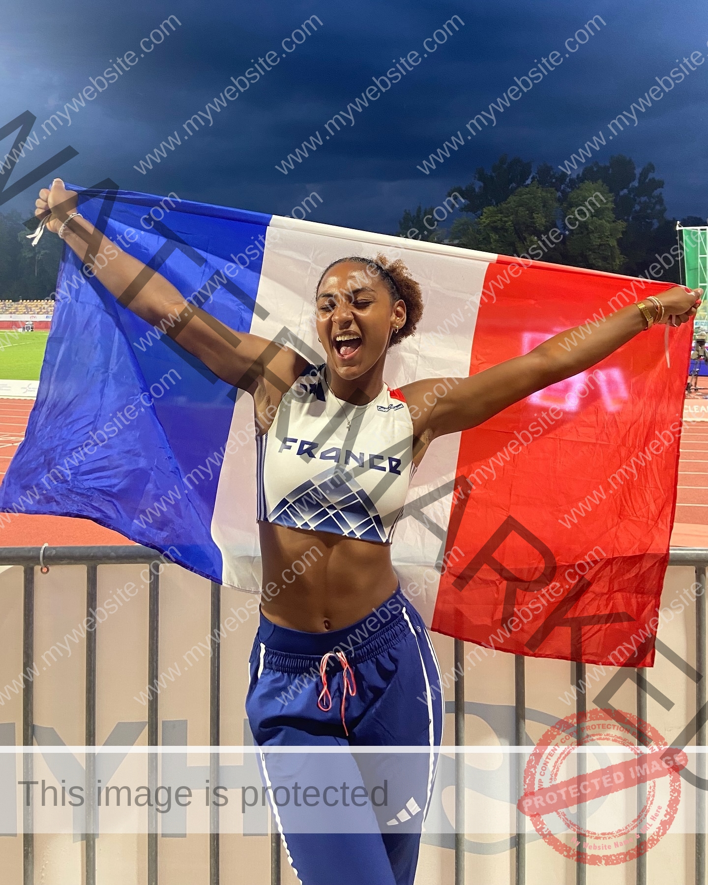 zola-ndouma-mona-france-zolaathle-09447 Zola Ndouma Mona, track and field star from France, joyfully holds a French flag at a stadium under a cloudy evening sky.