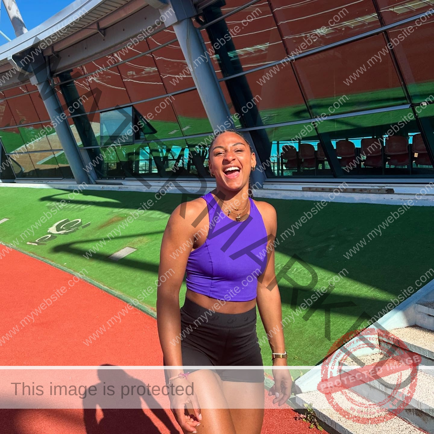 zola-ndouma-mona-france-zolaathle-09457 Zola Ndouma Mona, track and field star from France smiles and laughs in a purple top and black shorts on a running track by stairs.