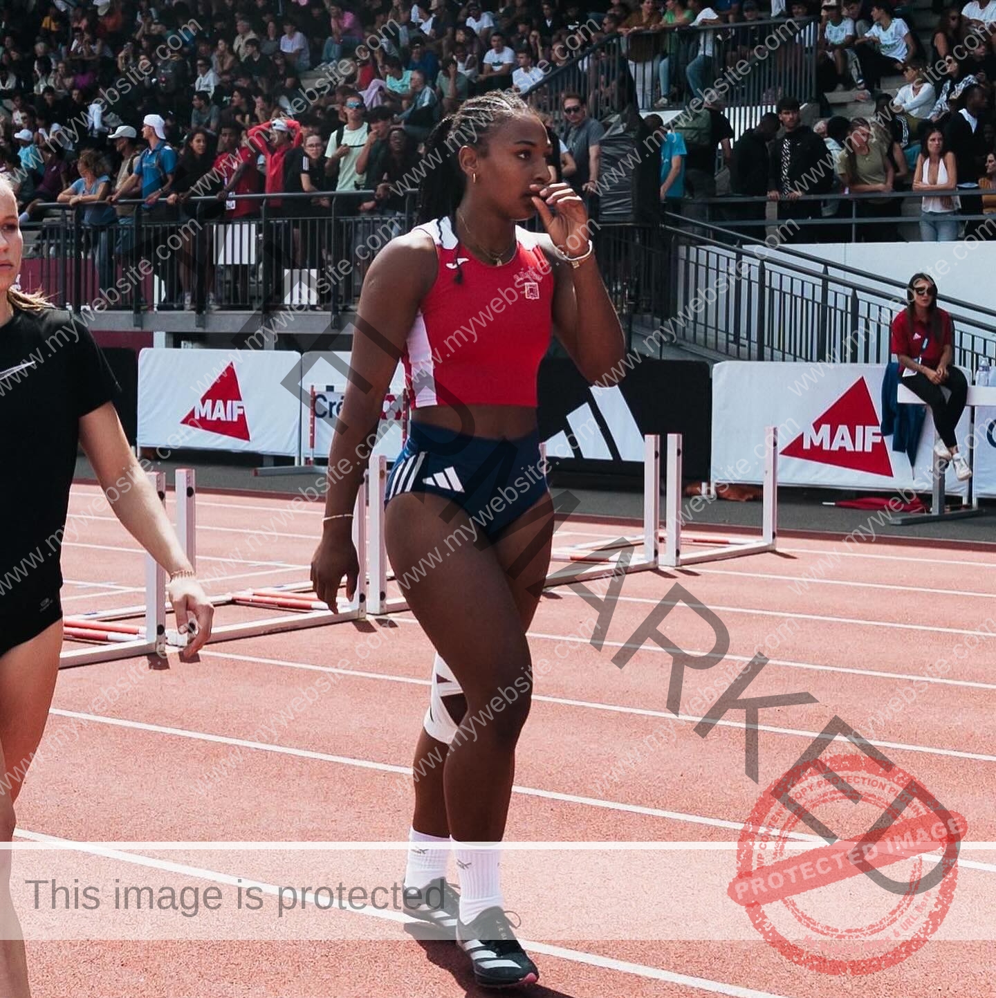 zola-ndouma-mona-france-zolaathle-09480 Zola Ndouma Mona, track and field star from France, stands thoughtful on the track in red and blue, with hurdles and a crowd behind.